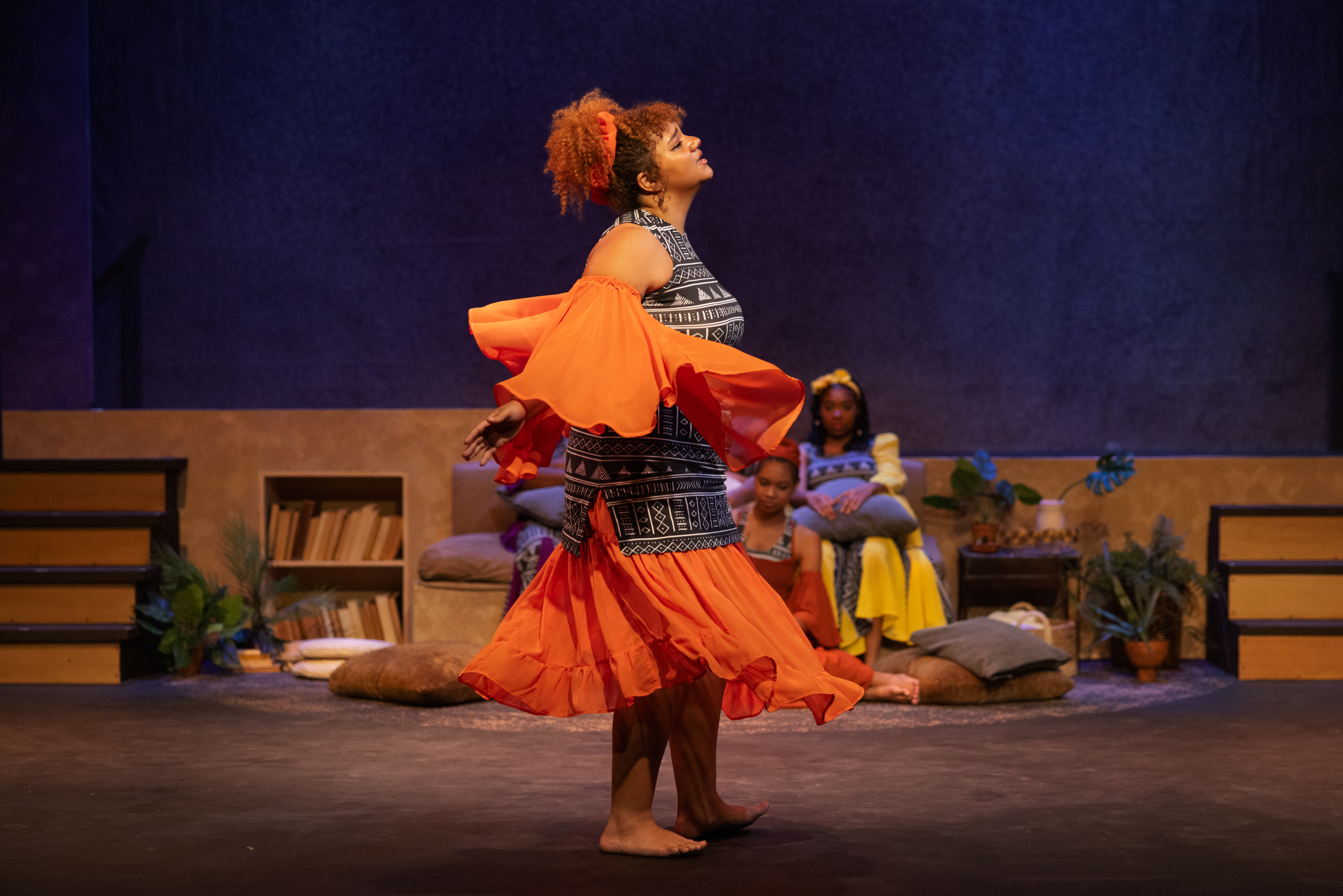 A Woman with curly hair tied up in an orange sash sways, the orange chiffon sleeves and skirt twirling around her