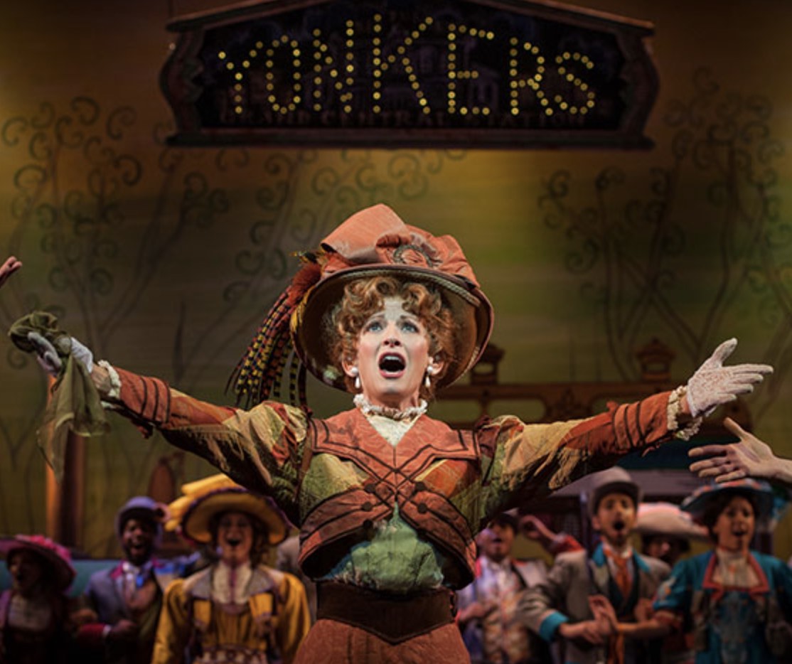 A woman holds her arms out with the palms up. She wears a large brimmed hat. Her mouth is open, holding a sung note.