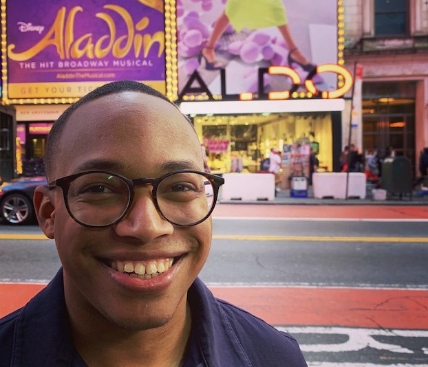 A young Black man wears black-rimmed glasses and smiles at the camera. He is standing in front of a billboard for ALADDIN the musical