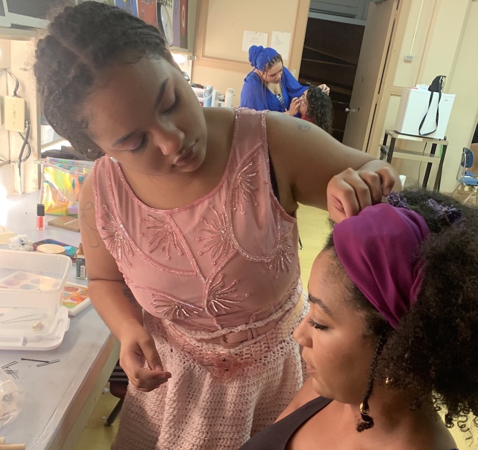 A young Black woman in a pink dress styles the hair of a young Black woman wearing a purple headband and sitting in a chair