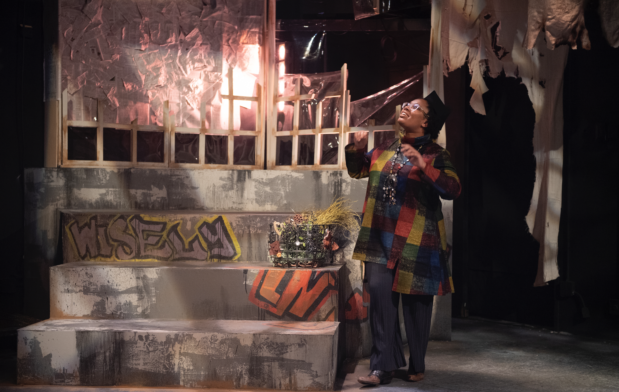 A young Black woman in a patchwork coat gazes up in a set filled with graffiti and trash