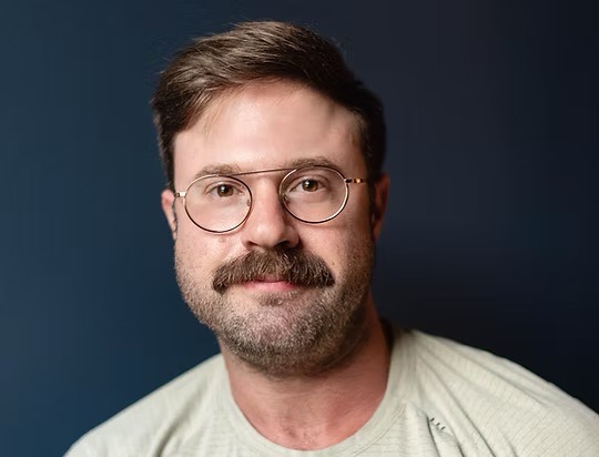 A white man with short hair, a mustache, beard, and glasses looks at the camera