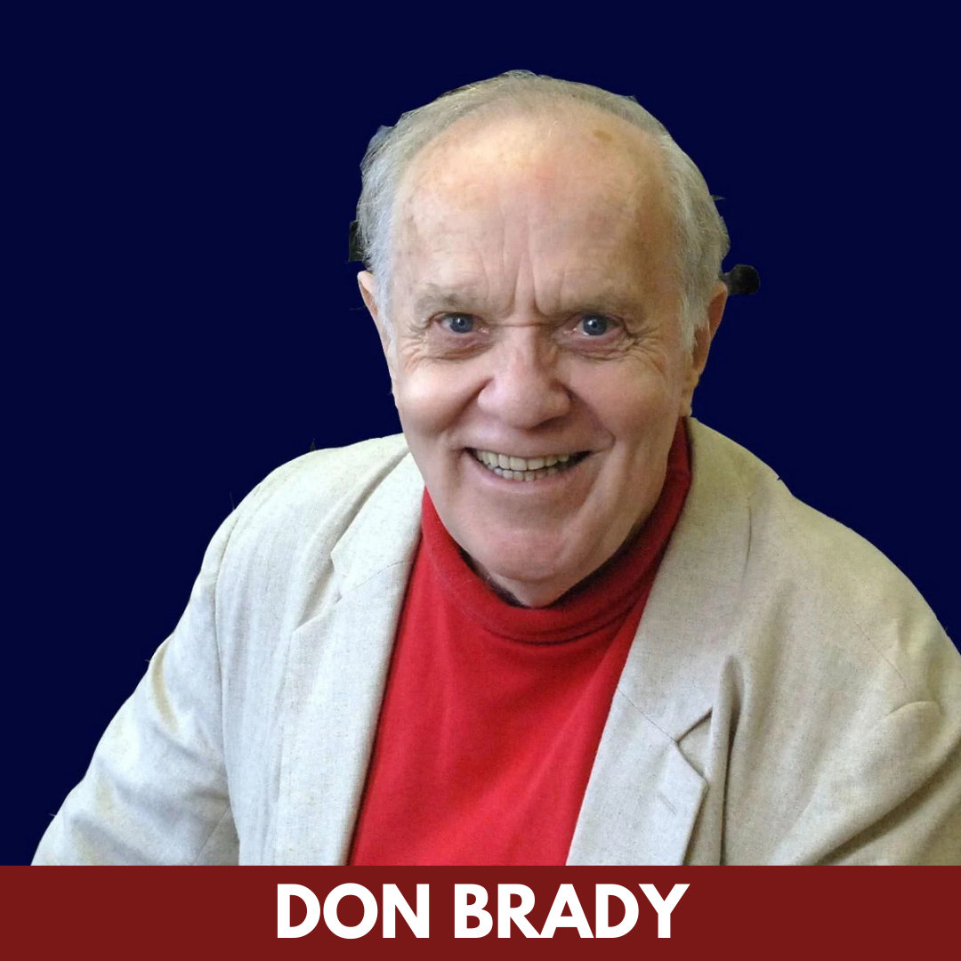 An older man with white hair smiles into the camera. He wears a tan suit jacket.