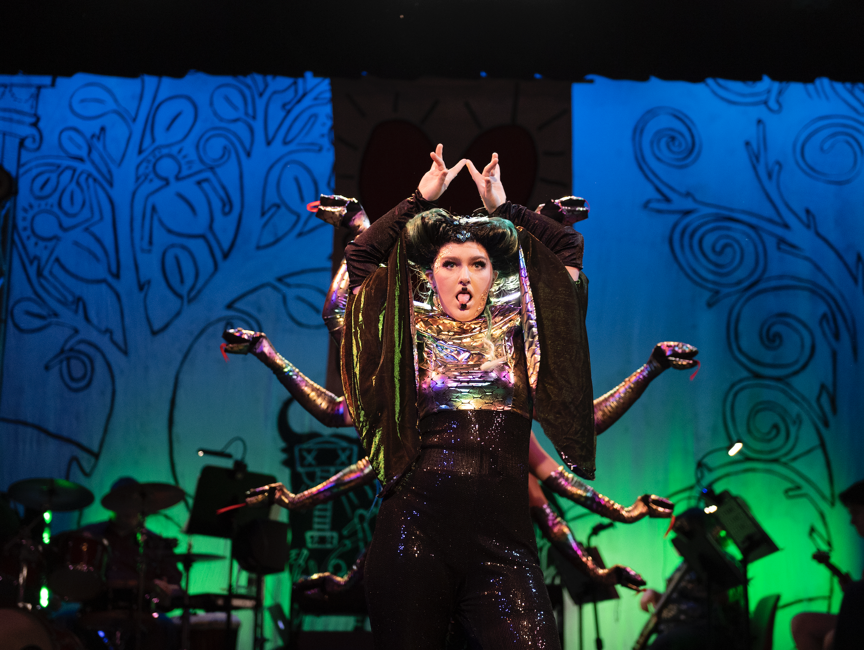 An actor wearing a shiny, metallic costume and raises their hands together over their head. Snake gloves surround them