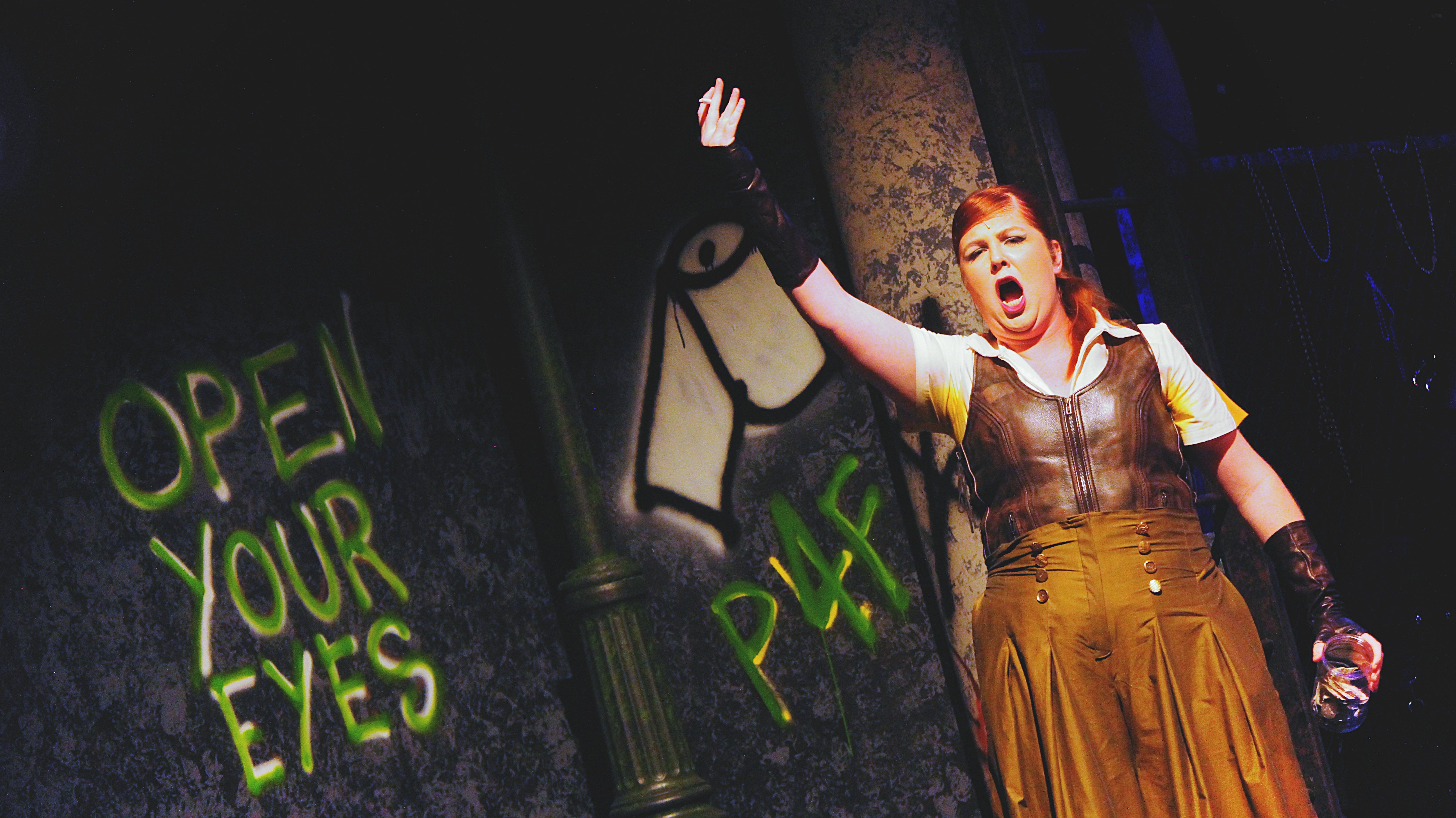 A tall red-headed woman in a dingy shirt and skirt raises her hand. Her mouth is open and she is singing!